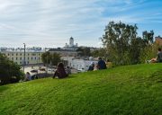 People relaxing on a grassy hill overlooking a city, with a white-domed cathedral and pale buildings in the distance under a blue sky.
