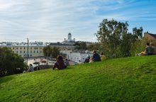 People relaxing on a grassy hill overlooking a city, with a white-domed cathedral and pale buildings in the distance under a blue sky.