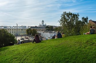 People relaxing on a grassy hill overlooking a city, with a white-domed cathedral and pale buildings in the distance under a blue sky.