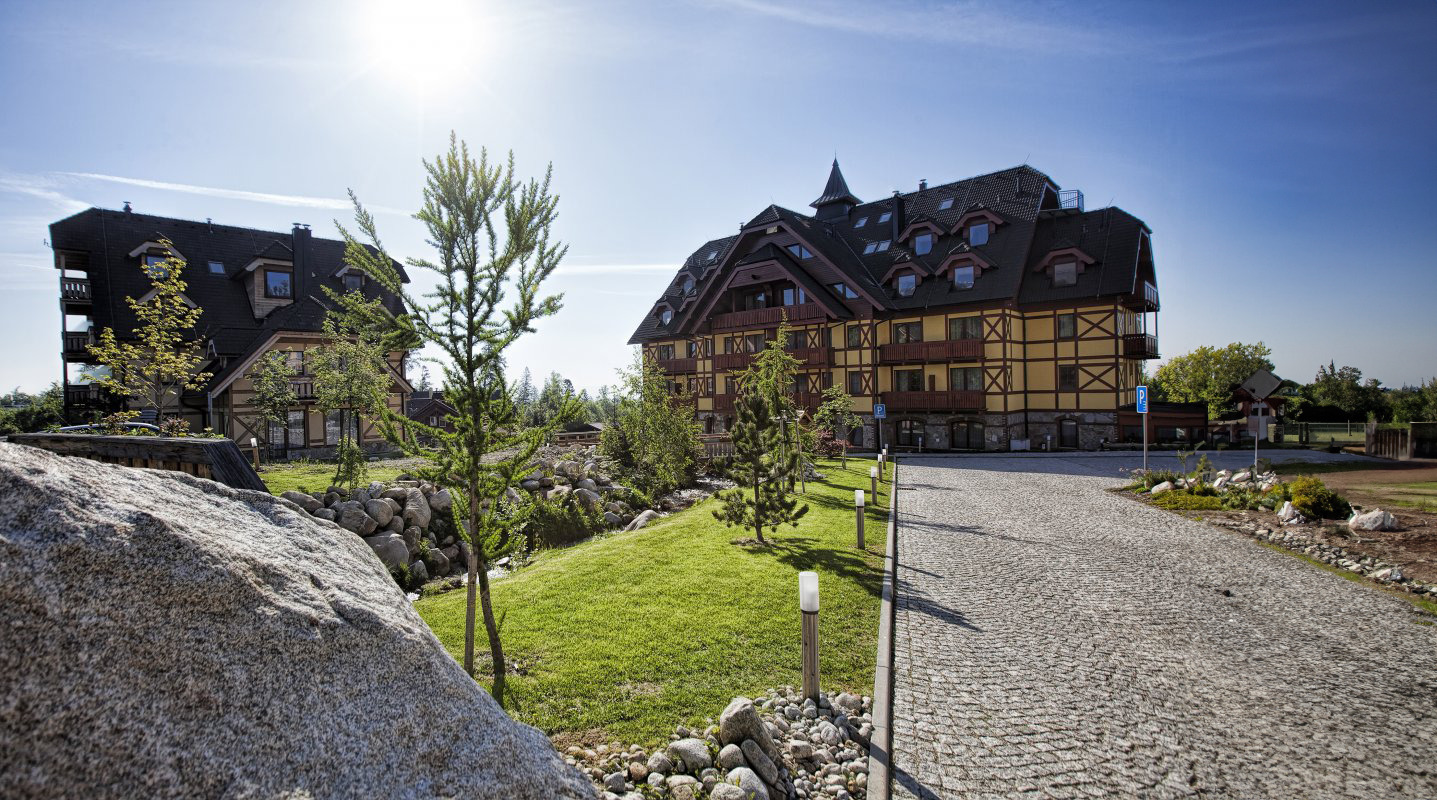 Chalet-style buildings along a cobblestone driveway with a green lawn, young trees, and rocks under a bright blue sky.