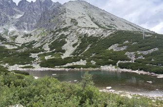 Alpine landscape with jagged rocky mountain, scree slopes, green shrubs, and a calm lake in the foreground; a cable car tower on the right side of the hill.