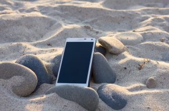 White smartphone partially buried in sand among smooth beach rocks on a sunny day, screen off.