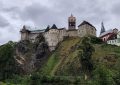 Castle perched on a rocky hillside under a cloudy sky, with a cluster of old buildings nearby and green vegetation. (informative)