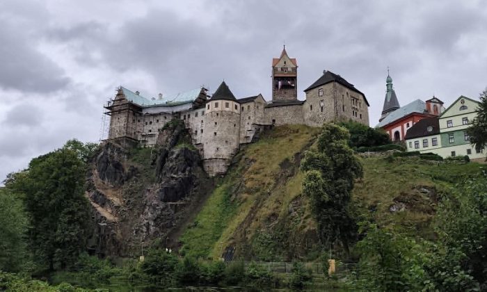 Castle perched on a rocky hillside under a cloudy sky, with a cluster of old buildings nearby and green vegetation. (informative)
