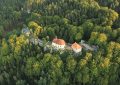 A white church with a red roof sits on a rocky cliff, surrounded by dense green forest from an aerial view.