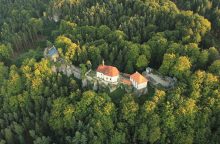 A white church with a red roof sits on a rocky cliff, surrounded by dense green forest from an aerial view.