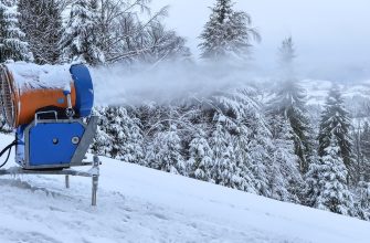 Snowmaking machine on a snowy hillside spraying artificial snow onto snow-covered trees in a misty forest landscape.