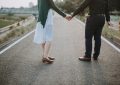 Couple seen from behind holding hands while walking down a deserted road, urban setting in the distance.