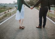 Couple seen from behind holding hands while walking down a deserted road, urban setting in the distance.
