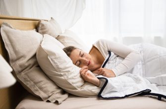 Woman sleeping on a bed with beige pillows and white quilt in a bright room, peacefully resting.